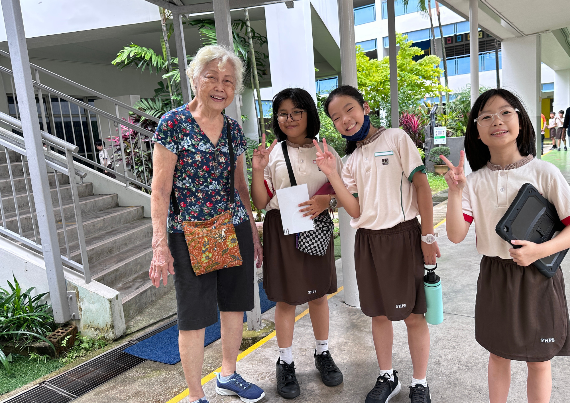 Elderly woman smiles beside three schoolgirls in uniforms giving peace signs in a covered outdoor walkway.